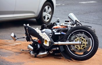 A motorcycle lies on its side on a roadway next to a white car, surrounded by sand.