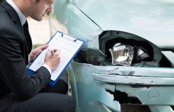 Man in suit writing on a clipboard while inspecting damage on the front bumper of a car.