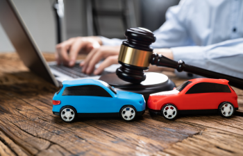 Toy cars colliding near a gavel on a wooden table, with a person typing on a laptop in the background.