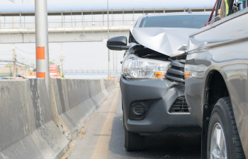 Two cars in a rear-end collision on a highway, with visible damage to the front of one vehicle. Concrete barriers line the road.