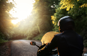 A person wearing a helmet rides a motorcycle on a tree-lined road with the sun shining in the background.