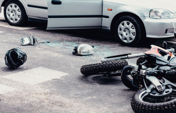 A car with an open door and a fallen motorcycle lie on the road. Debris, including a helmet and broken mirror, is scattered around.