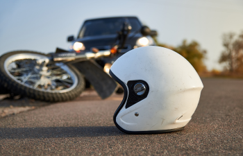 A white helmet lies on the road with a fallen motorcycle and a black car in the background.