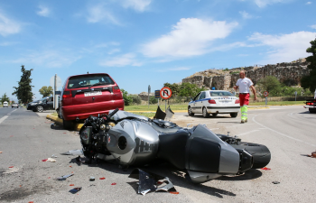 Motorbike lies on the road after a collision with a red car; a man in high-visibility clothing stands nearby; police car in the background.