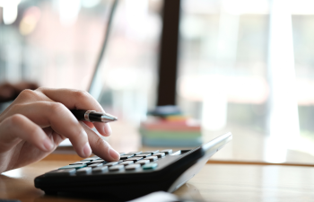 Person using a calculator with a pen in hand on a wooden desk. Blurred background with large windows.