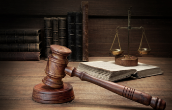 A wooden gavel rests beside an open book with scales of justice and stacked books in the background on a wooden table.