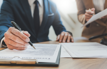 Two people in business attire at a desk, one holding a pen over a document on a clipboard, the other reviewing paperwork.