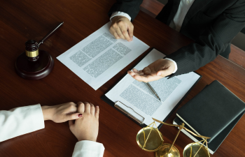 Two people in formal attire sit at a table with legal documents, a gavel, and scales. One person gestures towards the documents while the other listens with hands clasped.