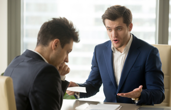 Two men in formal attire sit at a table in an office. One man gestures expressively while speaking, and the other listens intently with his hand on his chin.