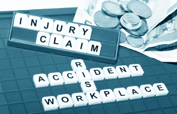 Scrabble tiles spell "Injury Claim," "Accident," "Workplace," and "Risks" on a textured surface. Nearby are scattered coins and banknotes.