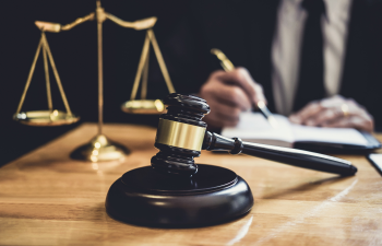 Gavel and scales on a desk with a person writing in the background, symbolizing legal proceedings.