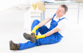 Man in blue overalls and yellow gloves sits on the floor, holding his foot, appearing to be in pain, in a bright indoor setting.