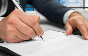 A person in a suit signing a document with a pen, against a backdrop of windows showing a cloudy sky.