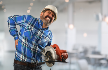 Man in a plaid shirt and hard hat holding his lower back in pain while carrying a circular saw.