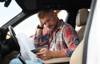 Man sitting in a car driver's seat, looking at his phone with a deployed airbag in front.