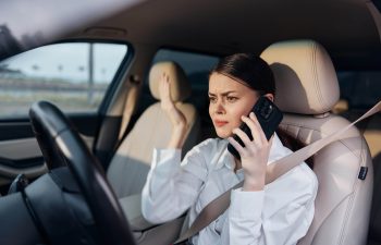 A woman in a car, wearing a seatbelt, holds a phone to her ear and raises her other hand with a frustrated expression.