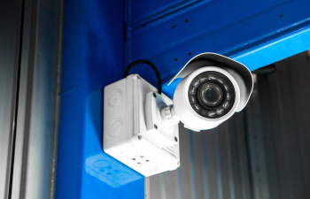 A white security camera is mounted on a blue wall corner. The camera has infrared LEDs surrounding the lens.