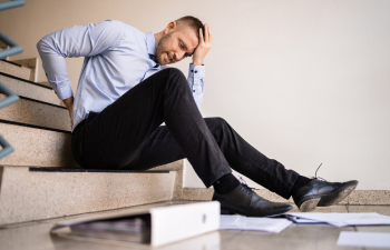 Man in a light blue shirt sits on stairs, holding his head and back in pain. Papers and a binder are scattered around him.