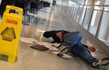 Person lying on the floor next to scattered papers in a hallway, holding their side. A caution sign is nearby, indicating a possible slip hazard.