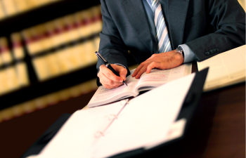 A person in a suit writes in an open book at a desk, surrounded by documents and shelves of books in the background.