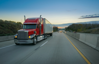 Red semi-truck driving on a highway during dusk, surrounded by trees on both sides, with a clear sky in the background.