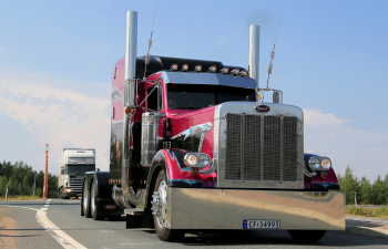 A large, shiny red and black semi-truck with chrome accents drives on a road. Another truck follows behind. Blue sky and trees are in the background.