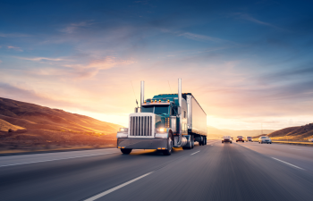 A large semi-truck drives on a multi-lane highway during sunset, with hills in the background and several cars nearby.