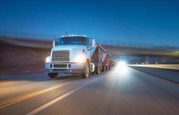 A white semi-truck moves rapidly on a highway at dusk, with blurred headlights in the background.