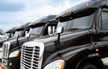 Close-up of a row of black semi-trucks parked in line, showcasing their front grilles and headlights under a cloudy sky.