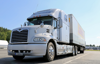 Silver semi-truck with a sleeper cab parked in a lot, trees and other vehicles in the background.