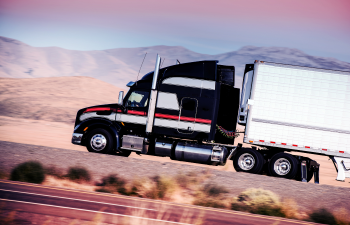 A black and red semi-truck with a white trailer drives on a highway through a desert landscape.