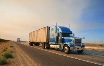 A large blue semi-truck with a trailer drives on a highway, followed by another truck, through a desert landscape under a clear sky.